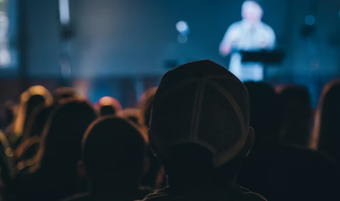 A Crowd Of People Listening To A Keynote Presentation